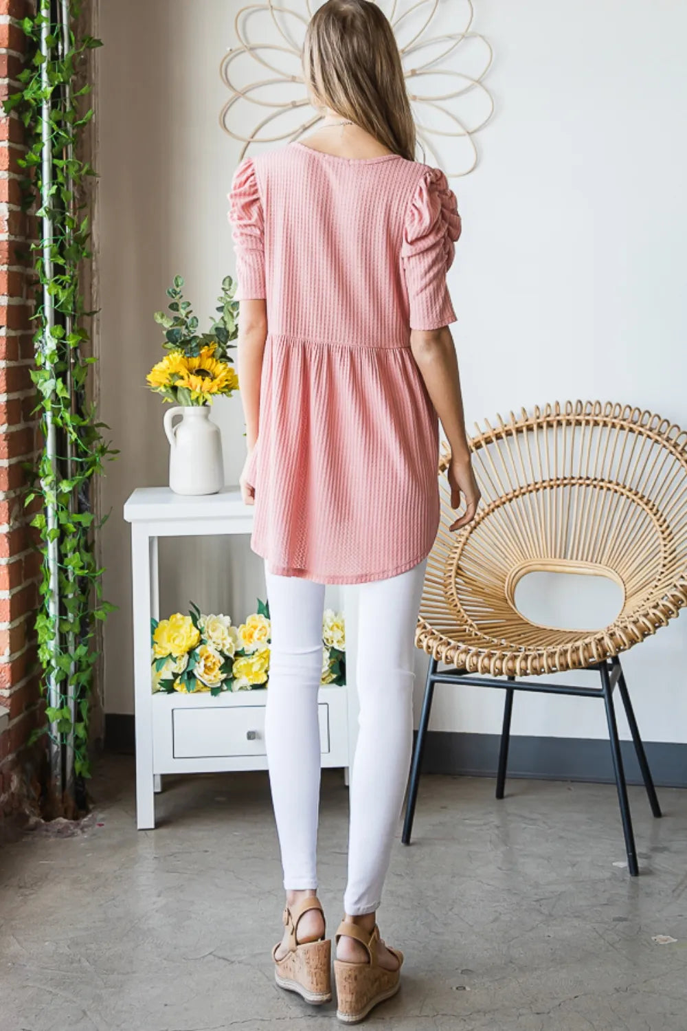 Woman wearing a pink dress and white pants standing in a room with a wicker chair and flowers.