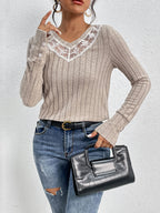 Woman wearing a beige ribbed sweater with lace neckline, blue jeans, and holding a black handbag against a light gray background.