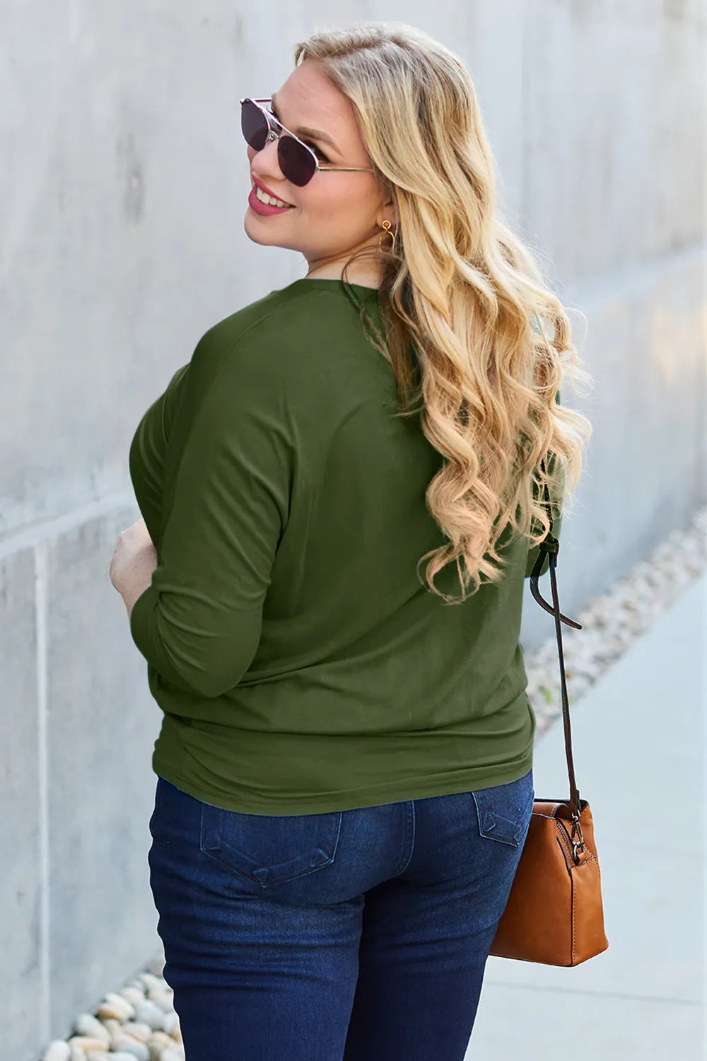 Woman wearing an olive green long-sleeve top and blue jeans, standing against a light-colored wall.