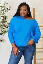 Woman wearing a blue sweater and jeans standing against a white background with a plant to the side.