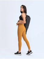 Woman wearing mustard yellow leggings and black sports bra with a gray bag over her shoulder on a white background
