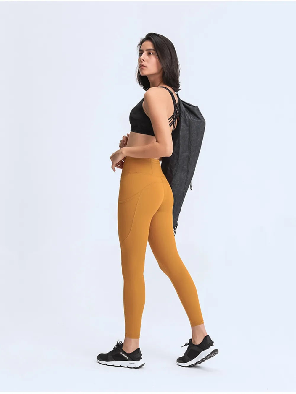 Woman wearing mustard yellow leggings and black sports bra with a gray bag over her shoulder on a white background