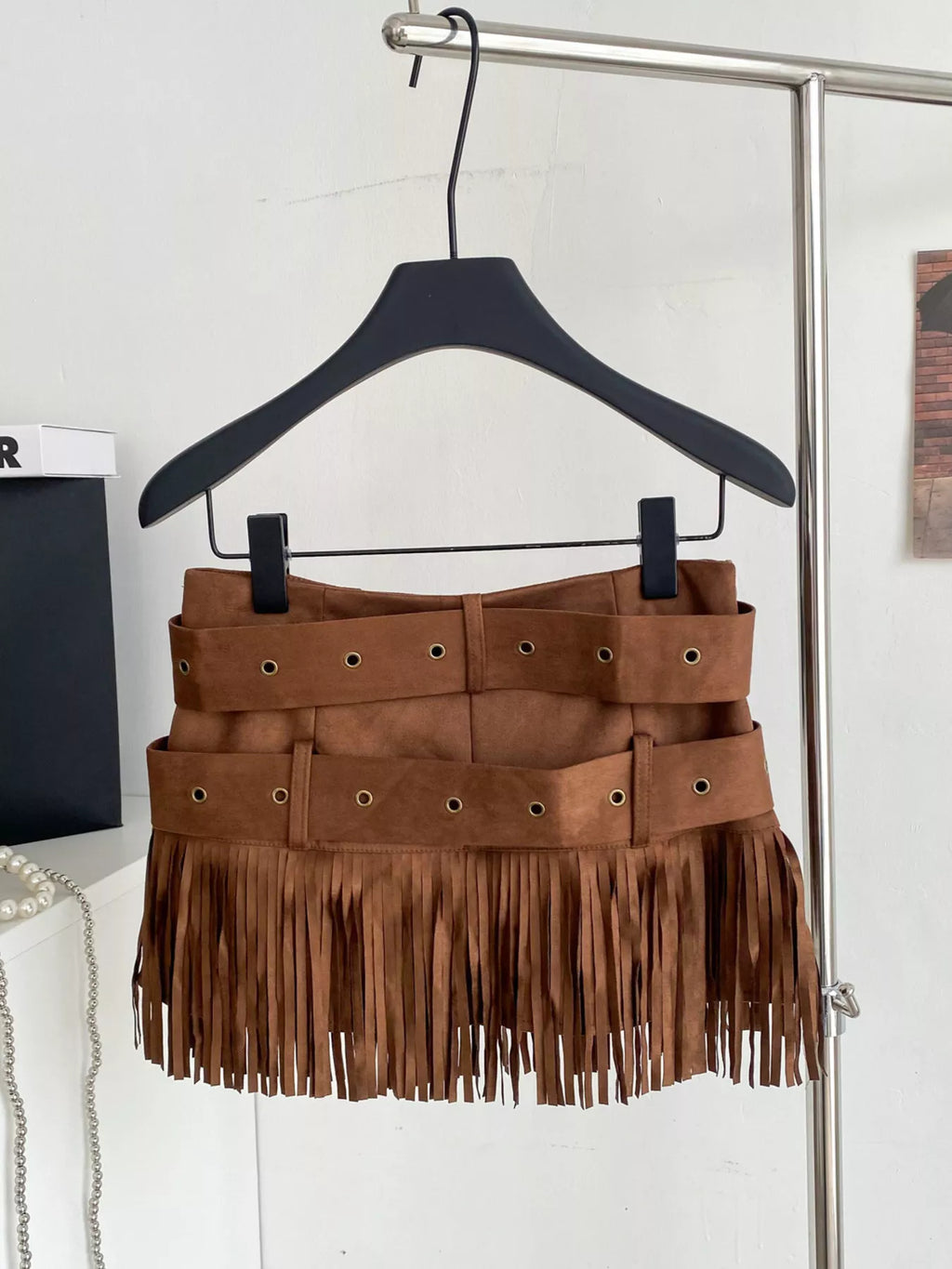 Brown suede fringe skirt on a hanger against a white wall.