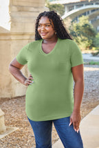 Woman wearing a green t-shirt and blue jeans standing outdoors with a stone wall and trees in the background.