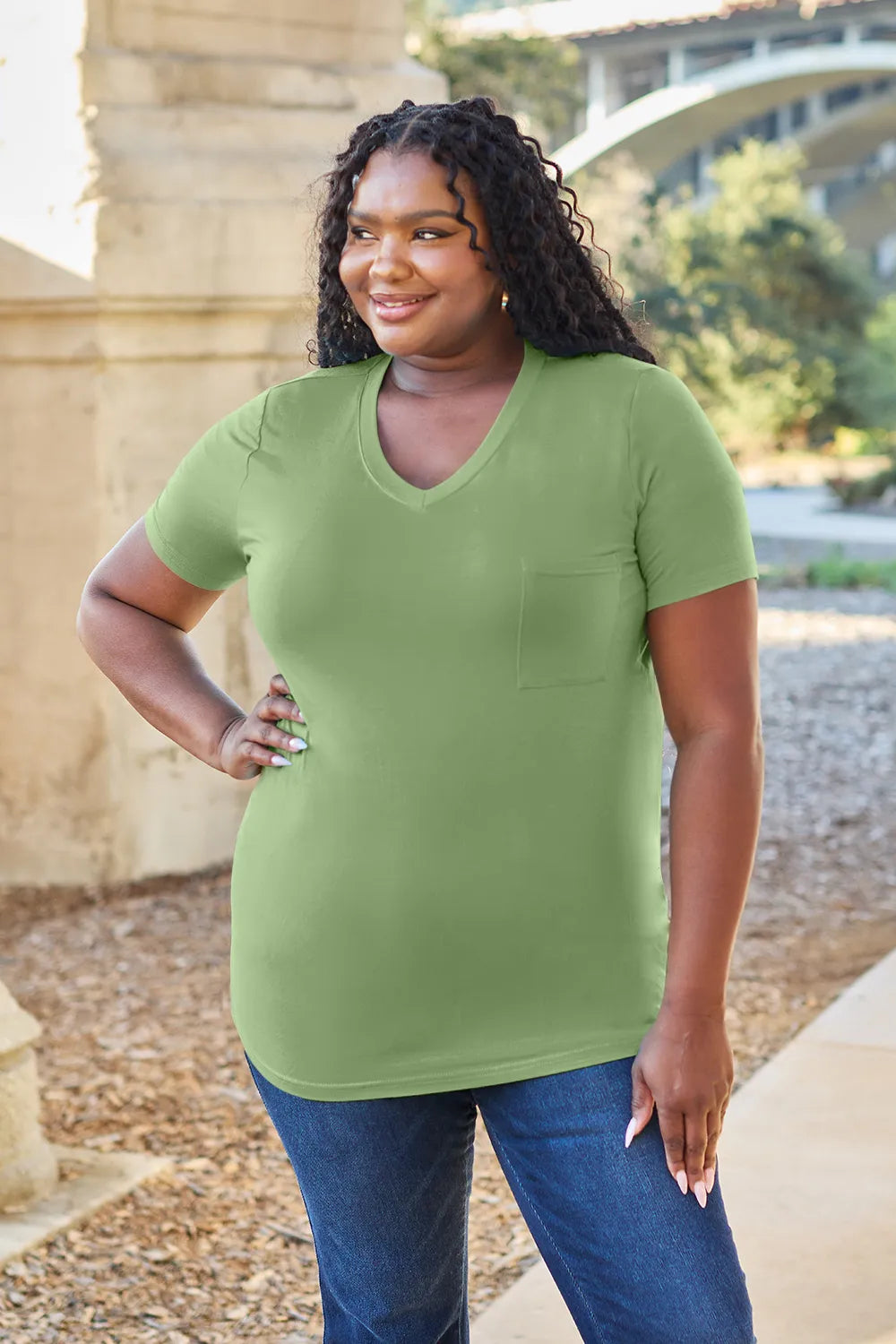 Woman wearing a green t-shirt and blue jeans standing outdoors with a stone wall and trees in the background.