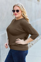 Woman wearing a brown top and blue jeans against a light gray wall.