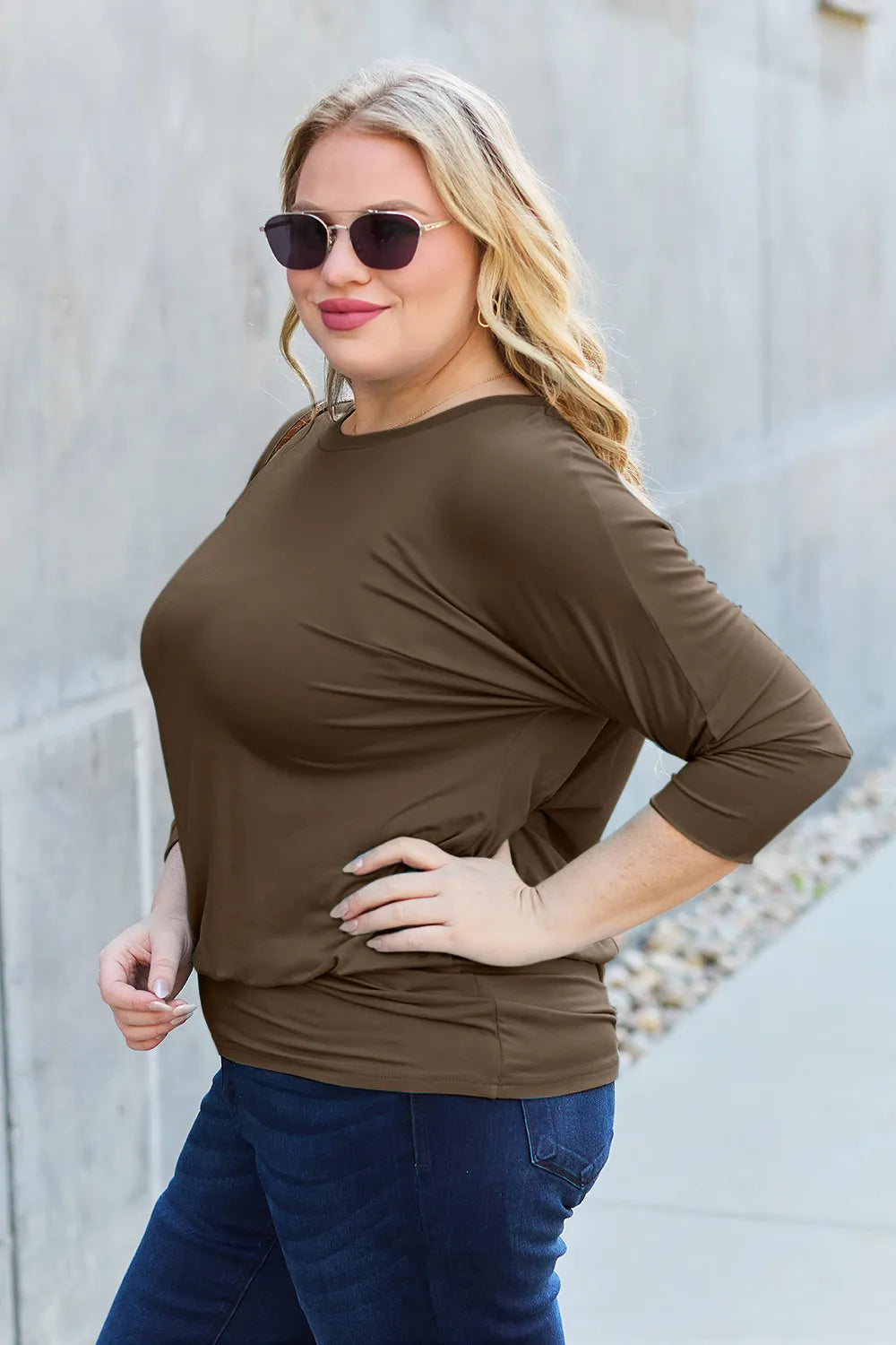 Woman wearing a brown top and blue jeans against a light gray wall.