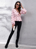 Woman wearing a pink blouse with butterfly patterns, black pants, and black boots against a gray wall.