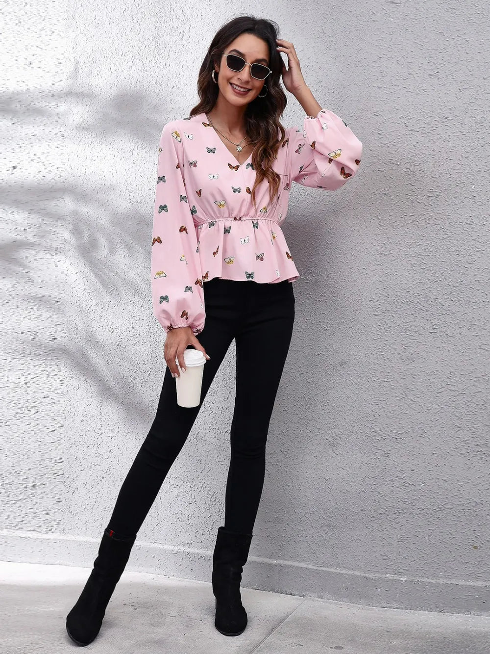 Woman wearing a pink blouse with butterfly patterns, black pants, and black boots against a gray wall.