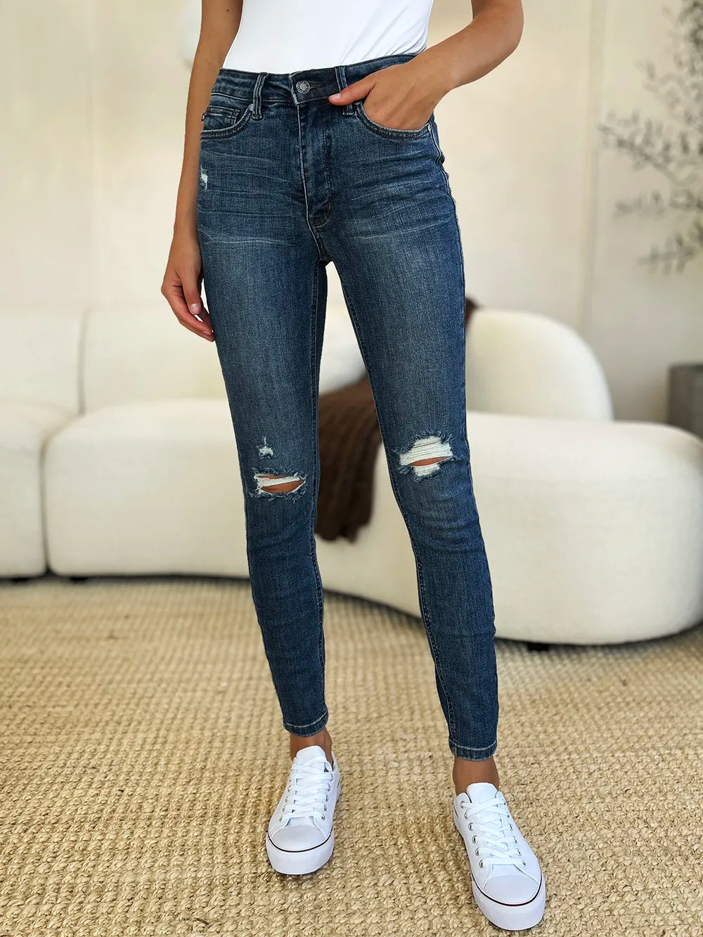 Person wearing blue jeans and white sneakers in a room with a beige sofa and carpet.
