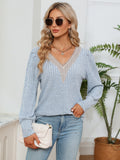 Woman wearing a light blue sweater and jeans, holding a white clutch, with a plant in the background.