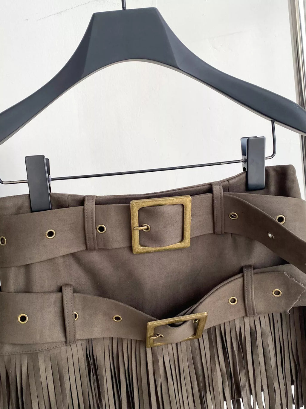 Brown fringe skirt with gold buckles on a hanger against a white background