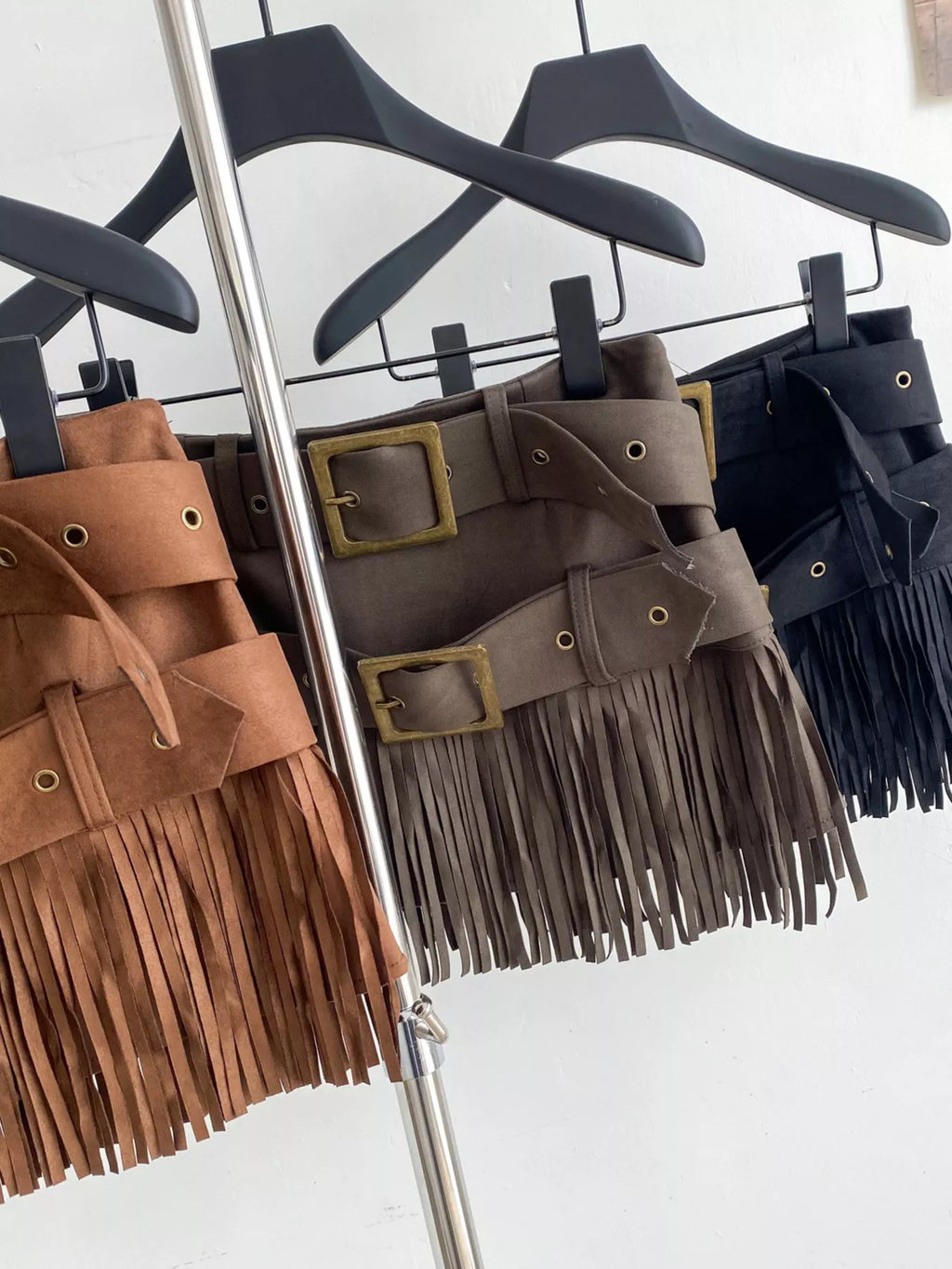 Three suede fringe belts in brown, gray, and black hanging on a rack.