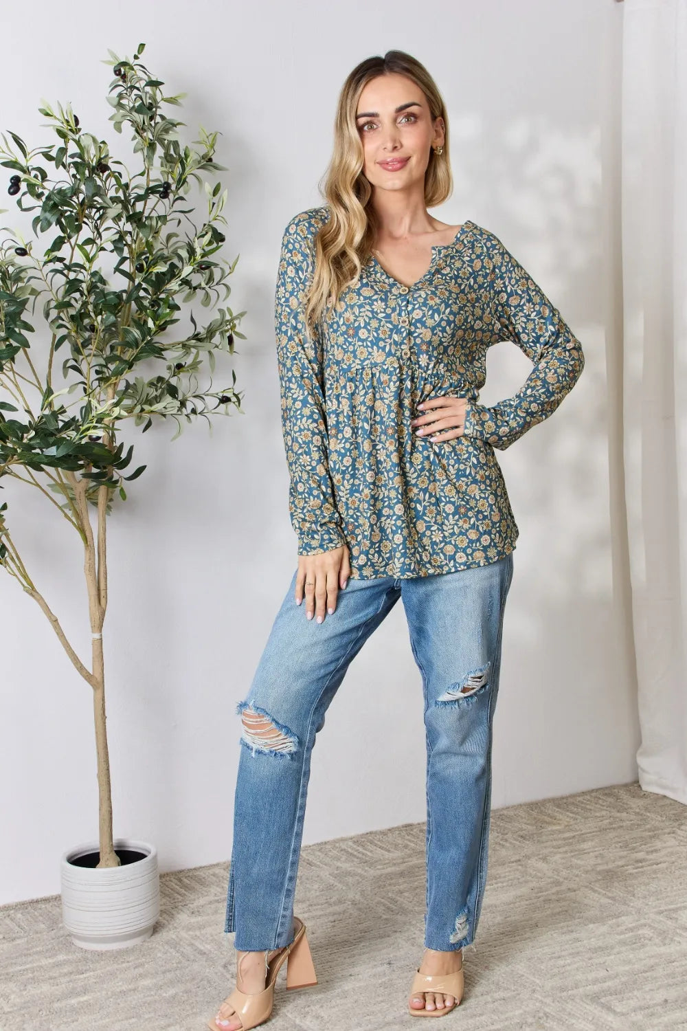 Woman wearing a floral long-sleeve top and ripped jeans standing next to a plant indoors.