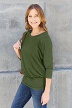 Woman wearing an olive green top and blue jeans against a concrete wall.