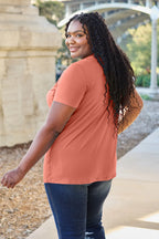 Woman wearing a rust-colored shirt and blue jeans standing outdoors.
