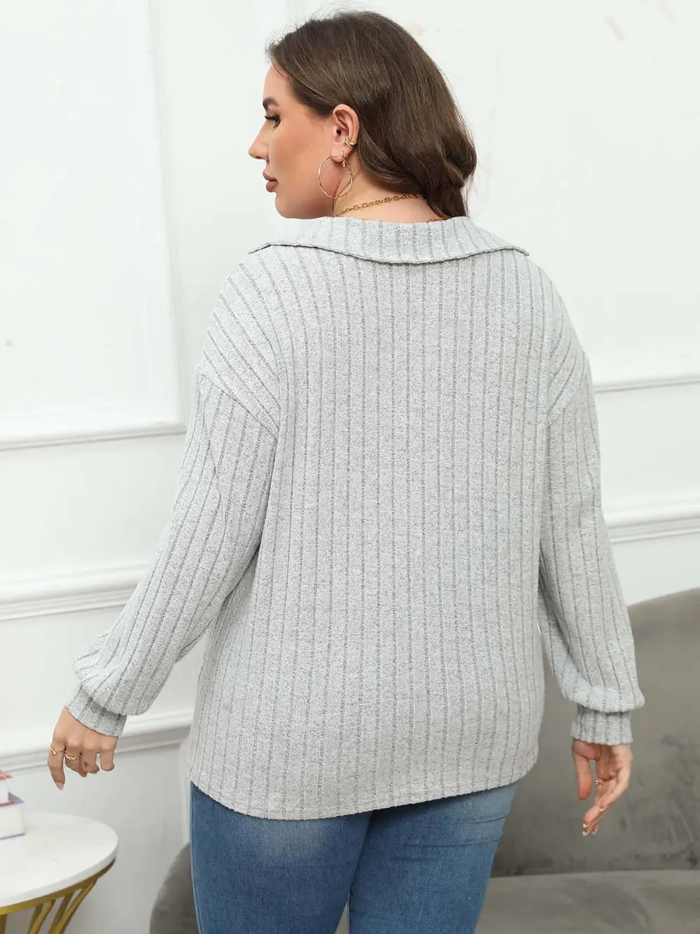 Woman wearing a light gray ribbed sweater and blue jeans in a minimal indoor setting.