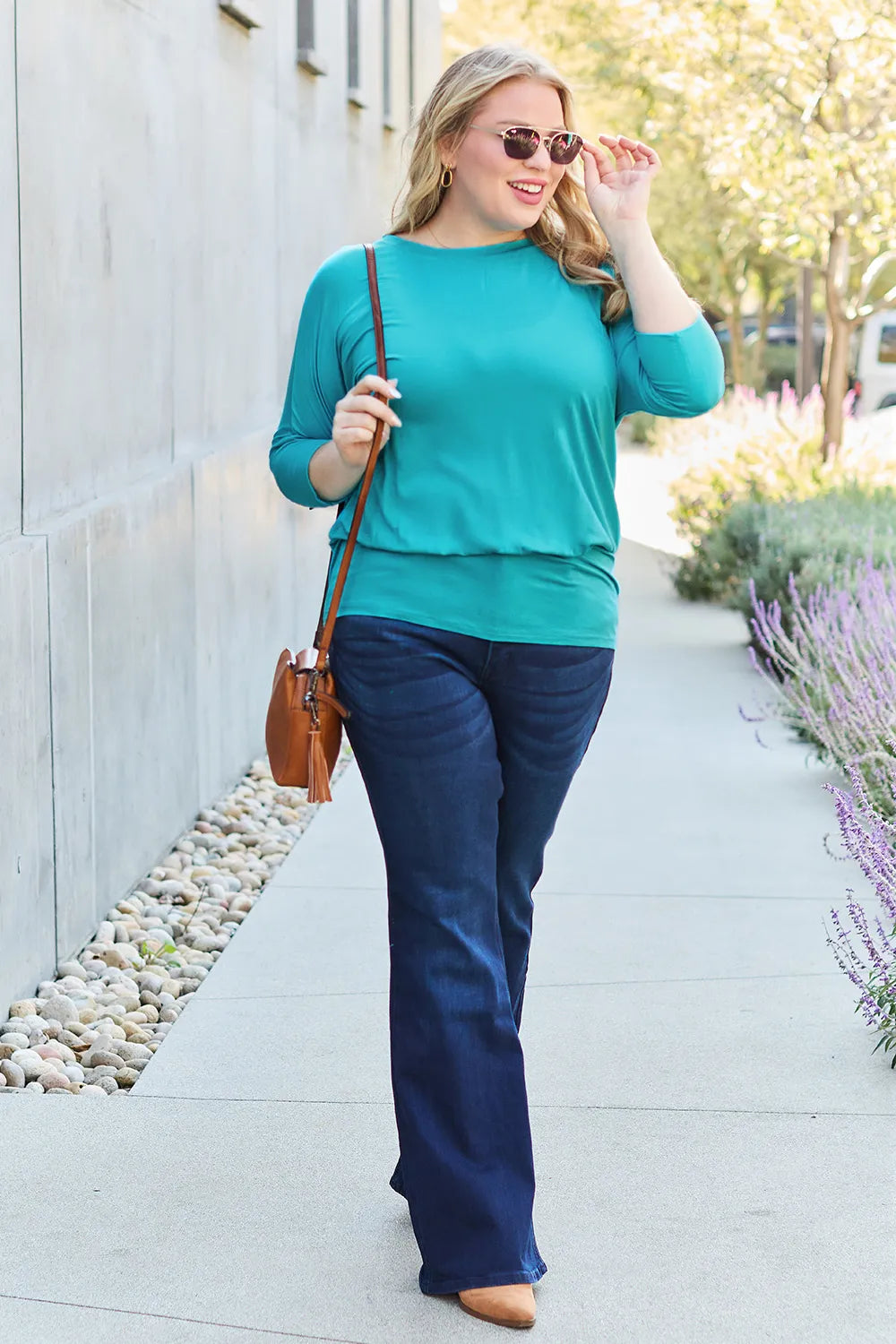 Woman in teal top and blue pants walking outdoors with a brown bag.