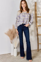 Woman wearing a floral blouse and dark jeans standing in a room with a white wall, wooden ladder, and decorative vase.