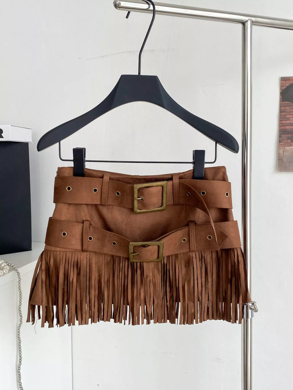 Brown suede skirt with fringe and buckles on a hanger against a white wall.