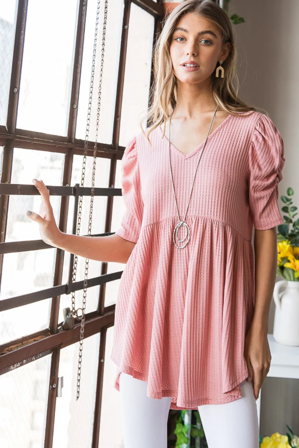 Woman wearing a pink blouse with a necklace in a room with a window and plants.