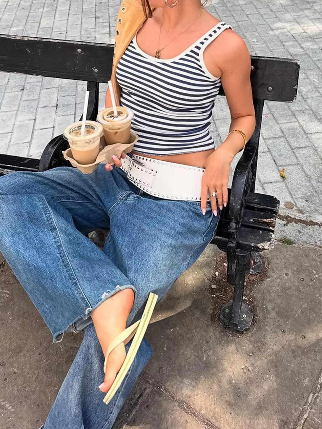Woman sitting on a bench holding iced drinks, wearing a striped top and jeans.