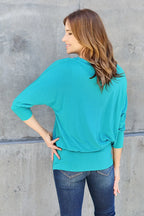 Woman wearing a turquoise top and jeans against a gray concrete wall