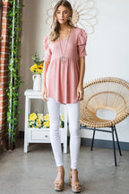 Woman wearing a pink top and white pants standing in a room with a wicker chair and flowers.