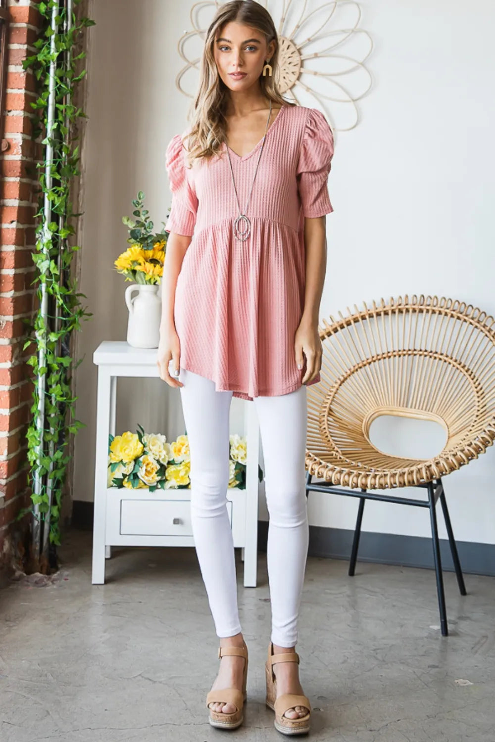 Woman wearing a pink top and white pants standing in a room with a wicker chair and flowers.