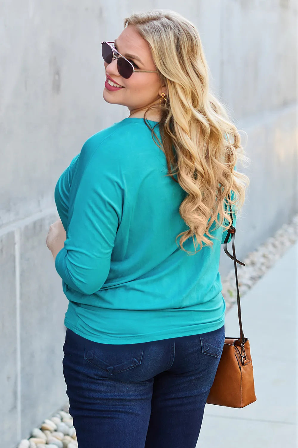 Woman wearing a teal long-sleeve top and blue jeans, standing against a light-colored wall.
