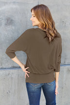 Woman wearing an olive green long-sleeve top and blue jeans against a gray concrete wall.