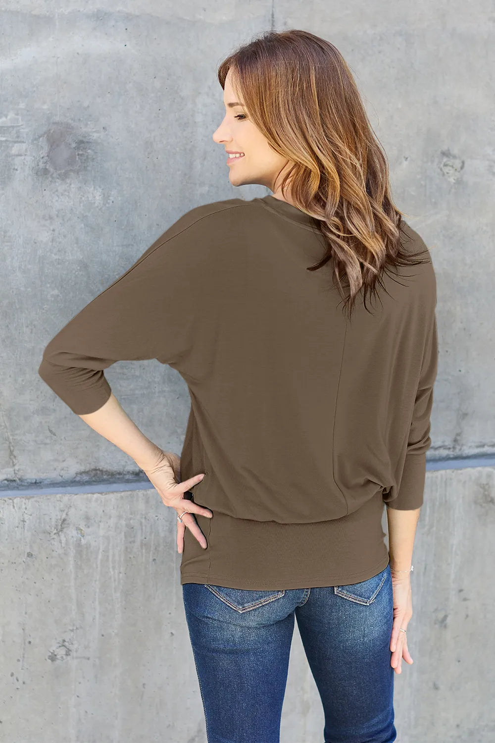Woman wearing an olive green long-sleeve top and blue jeans against a gray concrete wall.