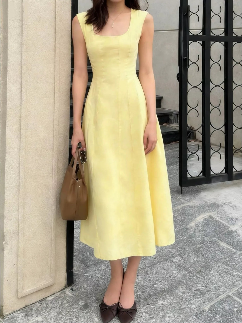 Woman wearing a yellow dress holding a brown handbag against a building exterior.