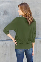 Woman wearing an olive green long-sleeve top and blue jeans against a gray concrete wall.