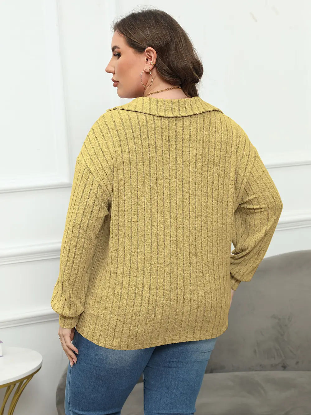 Woman wearing a mustard yellow ribbed sweater and blue jeans in a minimal indoor setting.