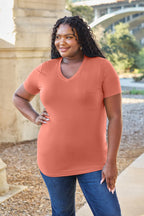 Woman wearing a terracotta-colored shirt and blue jeans standing outdoors with a bridge and trees in the background.