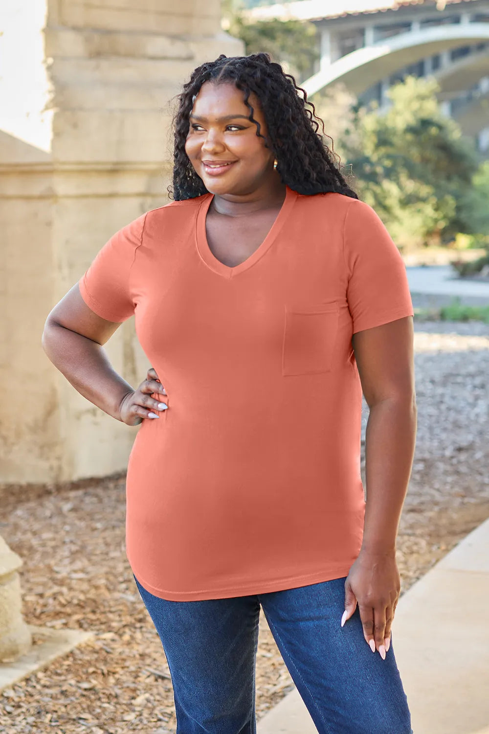 Woman wearing a terracotta-colored shirt and blue jeans standing outdoors with a bridge and trees in the background.