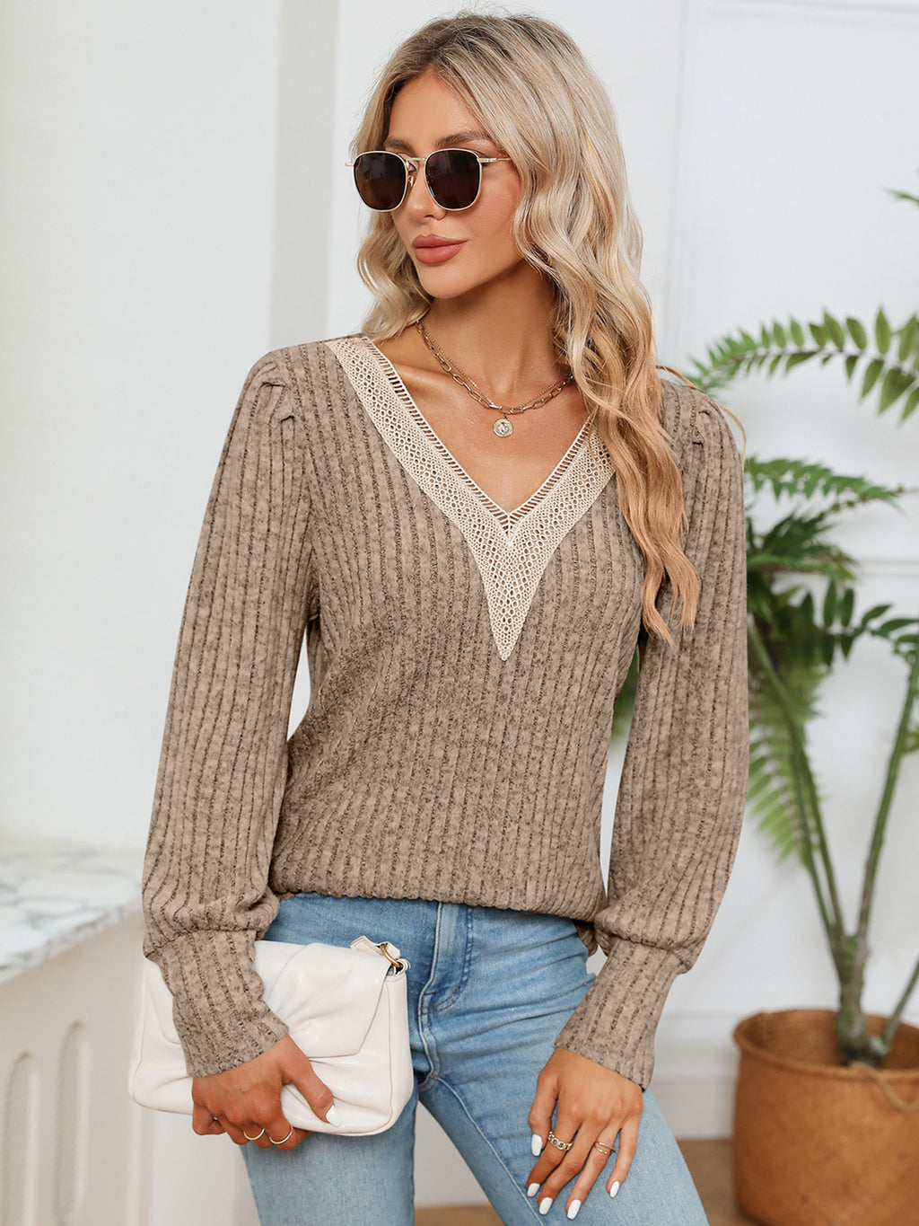 Woman wearing a brown ribbed sweater with lace neckline, holding a white clutch, standing indoors.