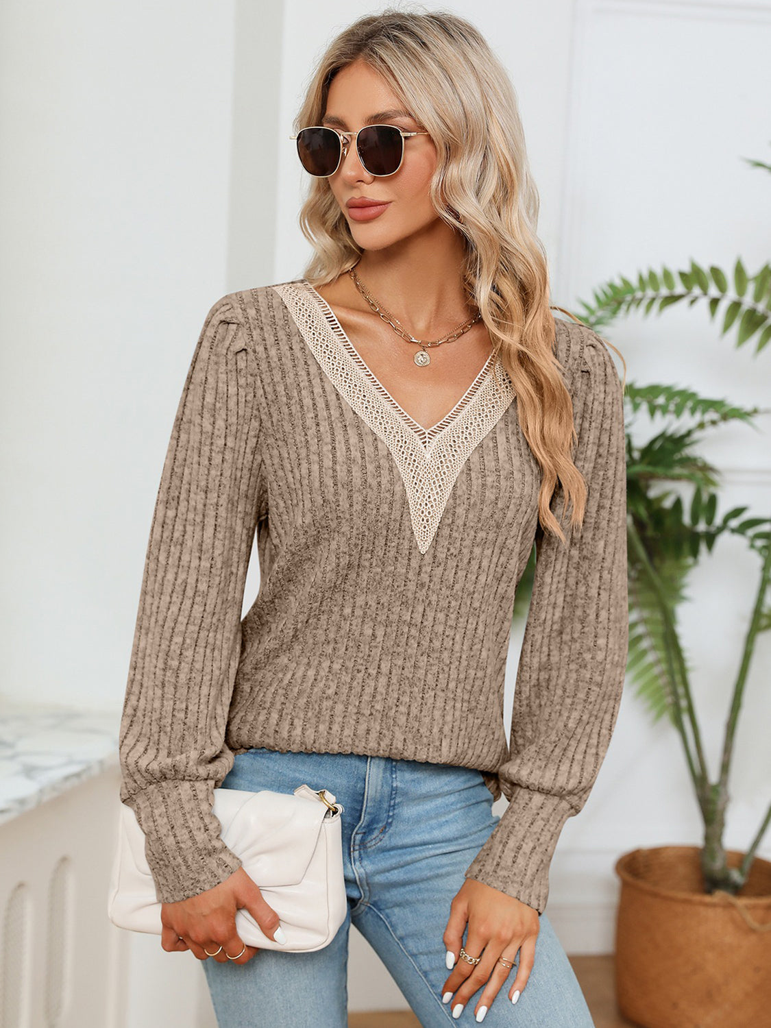 Woman wearing a brown ribbed sweater with lace neckline, holding a white clutch, standing indoors.