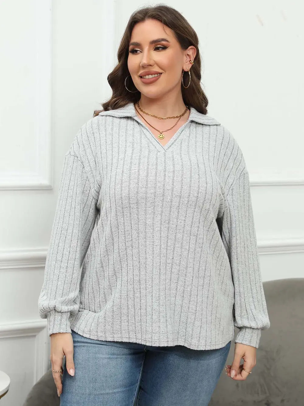 Woman wearing a light gray ribbed sweater with a hood and blue jeans against a white wall.