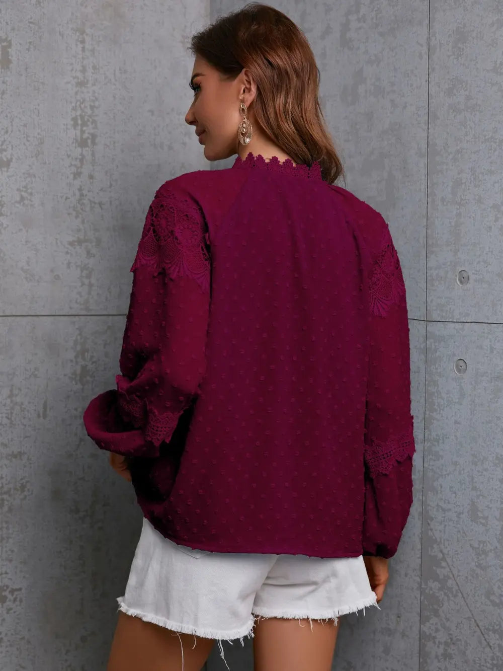 Woman wearing a burgundy blouse with lace details against a gray concrete wall.