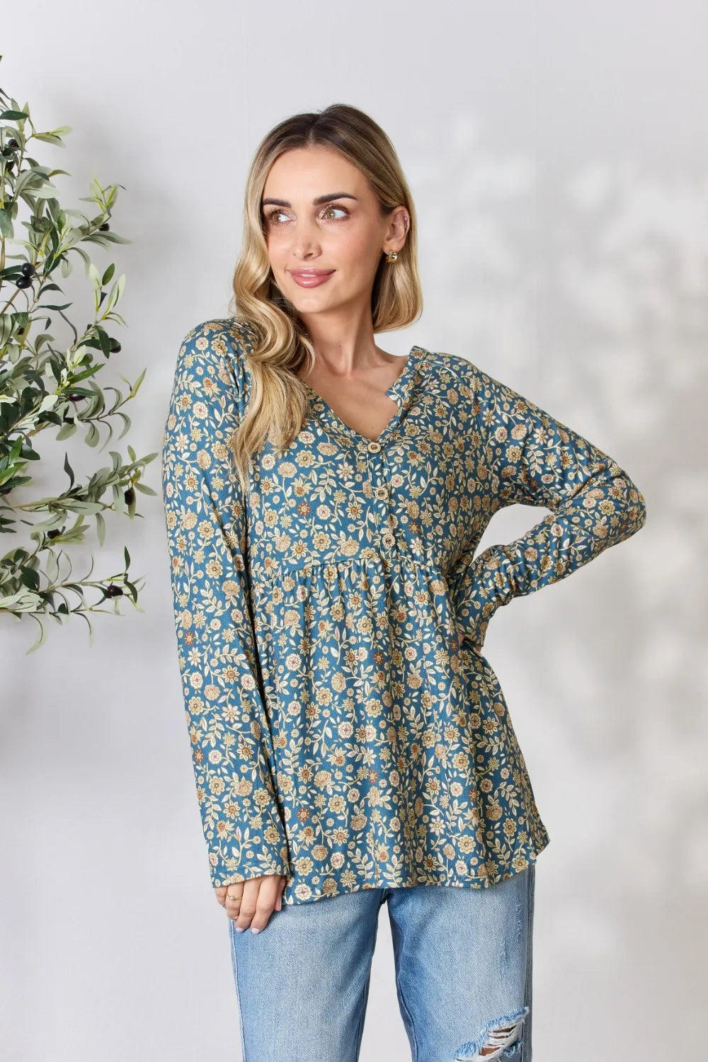 Woman wearing a floral blouse and jeans standing next to a plant on a light background