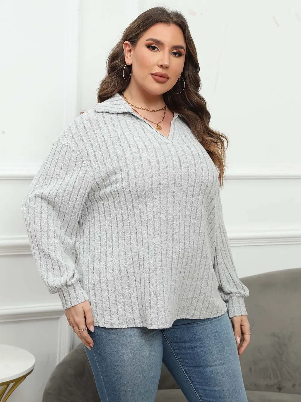 Woman wearing a light gray ribbed sweater and blue jeans indoors.