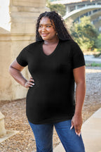 Woman wearing a black t-shirt and blue jeans standing outdoors with a stone building and greenery in the background.