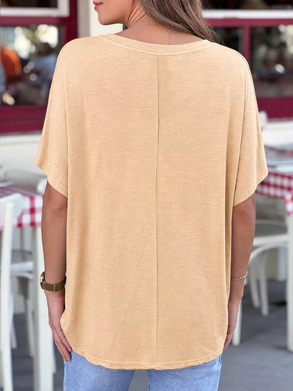 Person wearing a beige oversized t-shirt in an outdoor setting with tables and chairs.