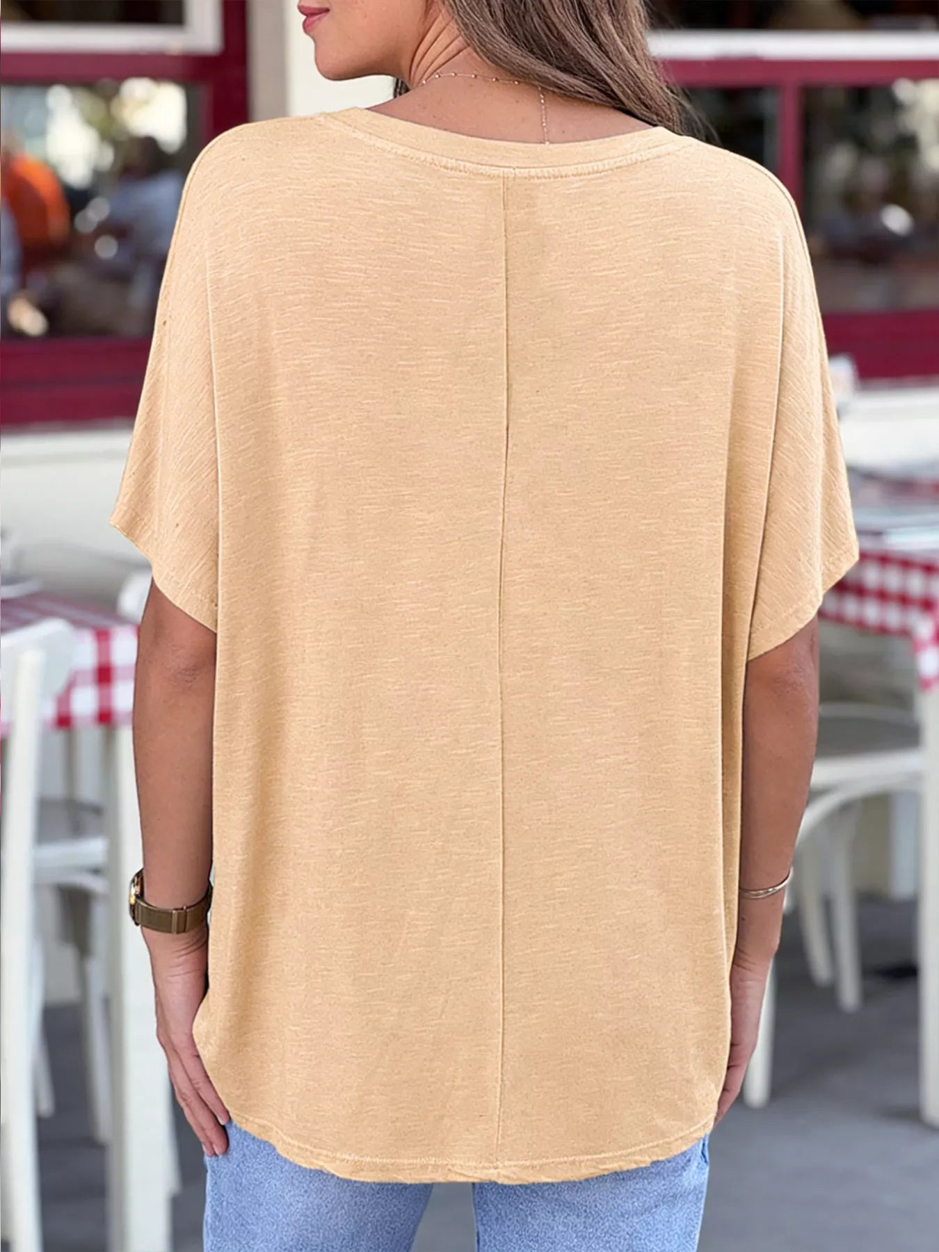 Person wearing a beige oversized t-shirt in an outdoor setting with tables and chairs.