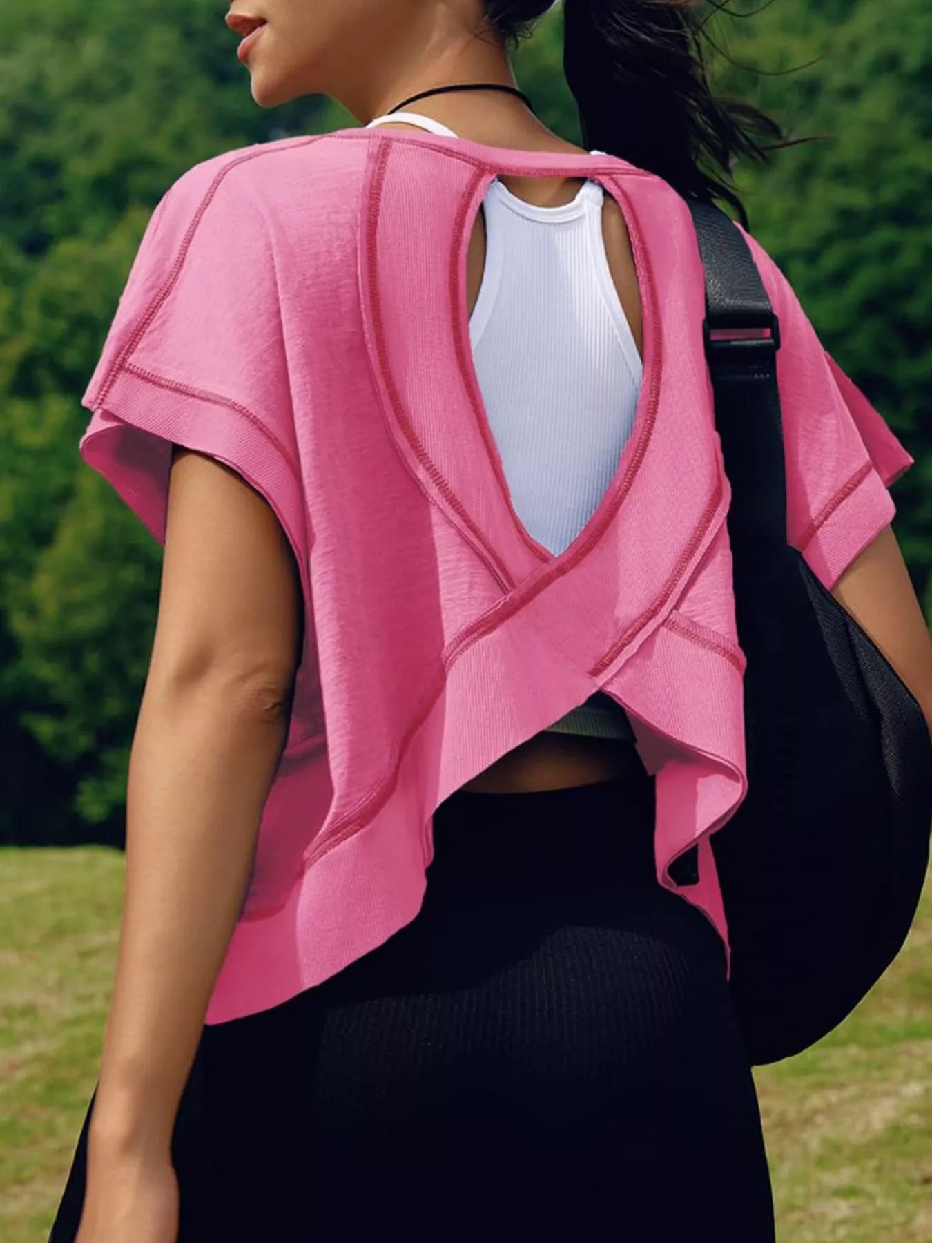 Person wearing a pink top with a white shirt underneath, standing outdoors with greenery in the background.