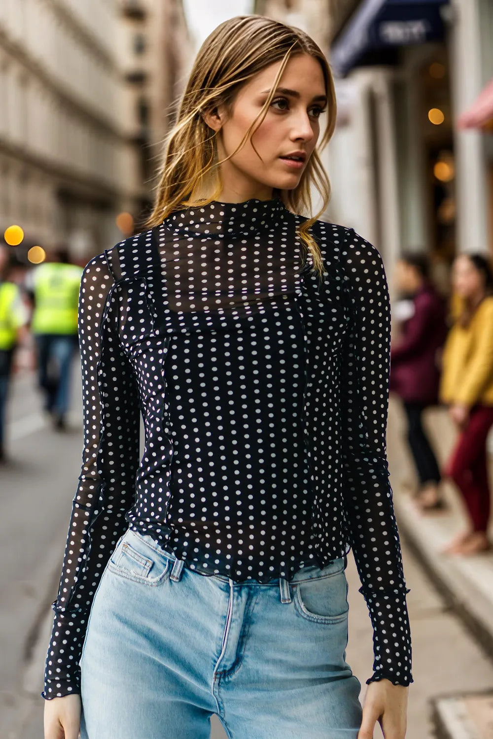 Woman wearing a black polka dot long-sleeve top and light blue jeans on a city street.