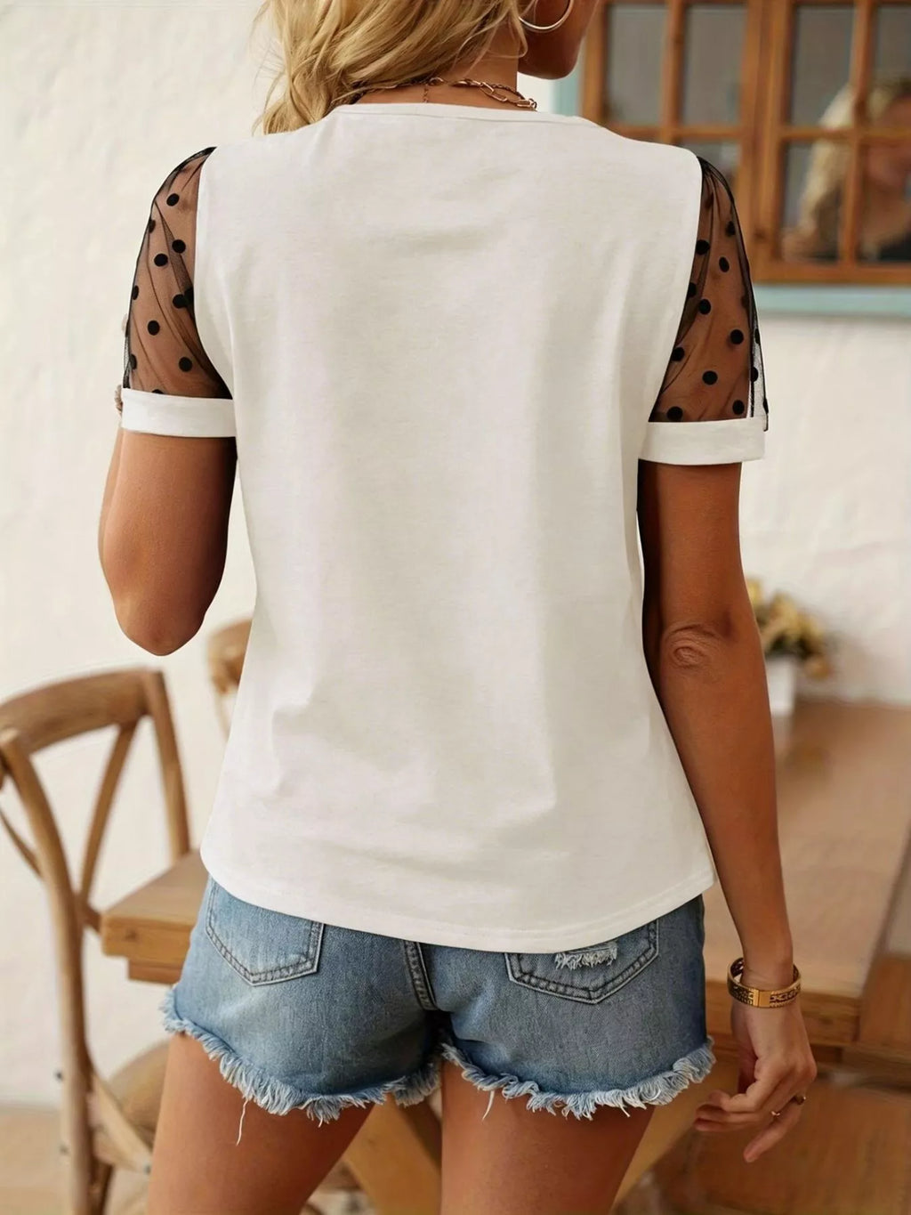 Woman wearing a white top with sheer sleeves and denim shorts in an indoor setting.