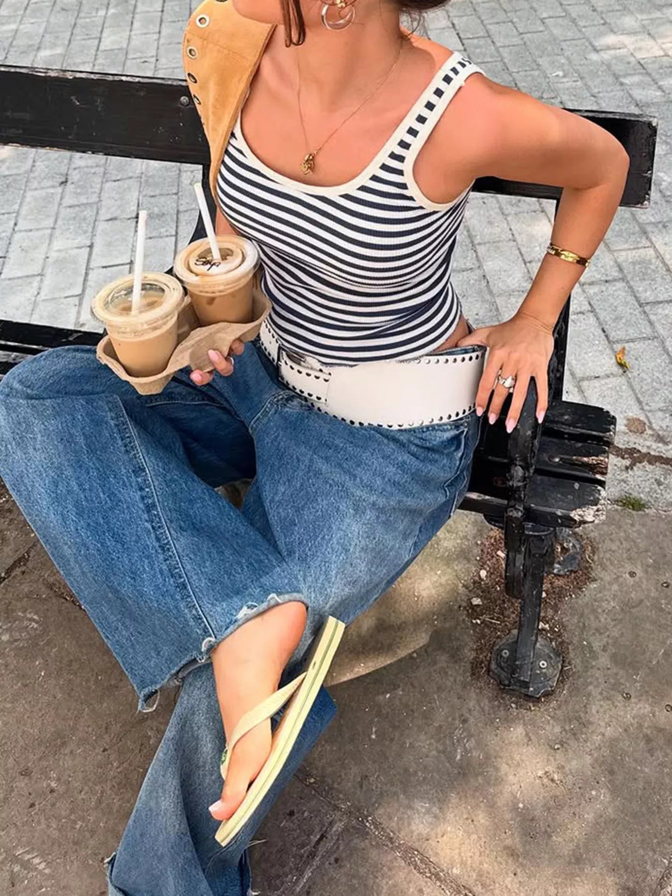 Woman sitting on a bench holding coffee cups, wearing a striped tank top and jeans.